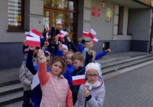 Grupa dzieci wychodzi na spacer z przedszkola. Dzieci wymachują swoimi flagami.
